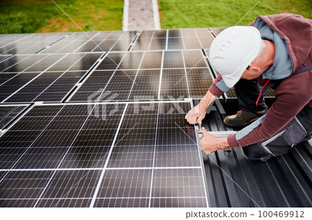 Man engineer mounting photovoltaic solar panels on roof of house. Technician in helmet installing solar module system with help of hex key. Concept of alternative, renewable energy. 100469912