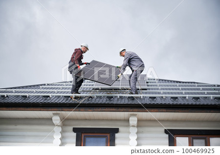 Mounters building solar panel system on roof of house. Men workers in helmets carrying photovoltaic solar module outdoors. Concept of alternative and renewable energy. 100469925