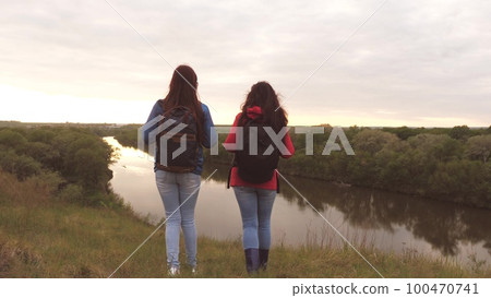 Girlfriends travel with backpacks by the river. Cheerful girls tourists are looking for adventure. Teamwork. Weekend on a sports hike among women. Rest at nature. Terrain research Girlfriends travel with backpacks by the river. Cheerful girls tourists are looking for adventure. Teamwork. Weekend on a sports hike among women. Rest at nature. Terrain research 100470741
