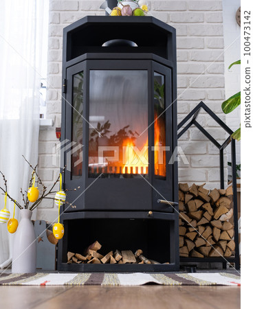 Easter decor near fireplace stove with fire and firewood. Cozy home hearth in interior with potted plans, colorful easter eggs, easter bunny and bouquet. Spring in a country house Easter decor near fireplace stove with fire and firewood. Cozy home hearth in interior with potted plans, colorful easter eggs, easter bunny and bouquet. Spring in a country house 100473142