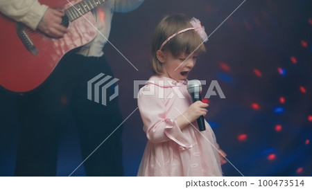 Little girl on stage in vintage dress, she sings into microphone and dances, her father plays acoustic guitar. Color music is shining and smoke is billowing. Father and Child Day, performance on stage 100473514