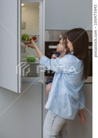 Mother holds a child in her arms and opens the refrigerator. 100475945