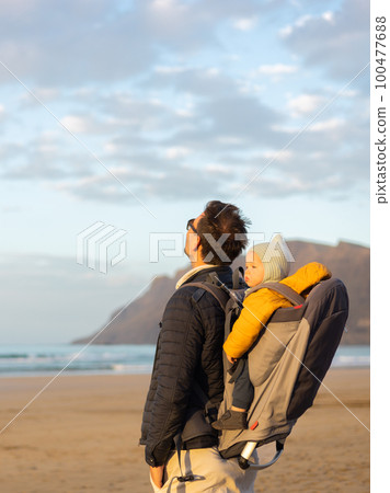 Father rising hands to the sky while enjoying pure nature carrying his infant baby boy son in backpack on windy sandy beach of Famara, Lanzarote island, Spain at sunset. Family travel concept 100477688