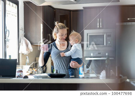 Happy mother holding her little infant baby boy while drinking morning coffee and making pancakes for breakfast in domestic kitchen. Family lifestyle, domestic life concept 100477698