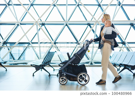 Mother carying his infant baby boy child, pushing stroller at airport departure terminal moving to boarding gates to board an airplane. Family travel with baby concept 100480966