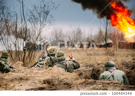 Fight Against Combat Vehicle. Re-enactors Armed Rifles And Dressed As World War Ii German Wehrmacht Infantry Soldiers Fighting Defensively In Trench. Building On Fire On Background 100484349