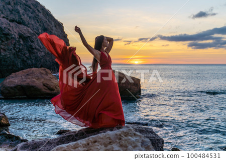 Woman in a red flying dress on the ocean or on the sea beach against the backdrop of the sunset sky. Woman in a red flying dress on the ocean or on the sea beach against the backdrop of the sunset sky. 100484531