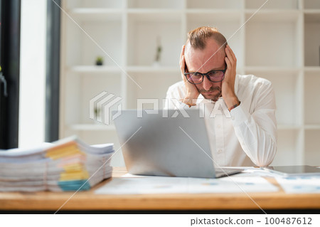 Tired businessman looking at laptop while sitting at desk in office Tired businessman looking at laptop while sitting at desk in office 100487612