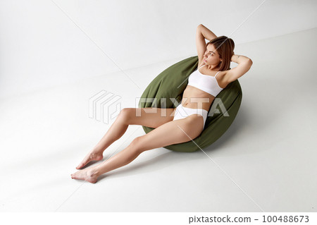 Beautiful woman sitting on big pillow chair in white underwear isolated over grey background. Sportive body. Concept of beauty, skin care, health, cosmetics, relax time 100488673