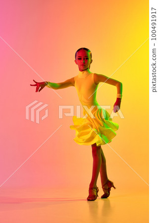 Expressive little girl in adorable stage outfit, dress dancing ballroom dance over gradient pink-yellow background in neon light filter. Concept of beauty, professional dances, skills 100491917