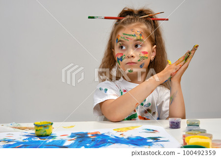 Little girl in white t-shirt, with brushes in her hair is sitting at the table with whatman and paints, painting on it. Isolated on white. Close-up. Little girl in white t-shirt, with brushes in her hair is sitting at the table with whatman and paints, painting on it. Isolated on white. Close-up. 100492359