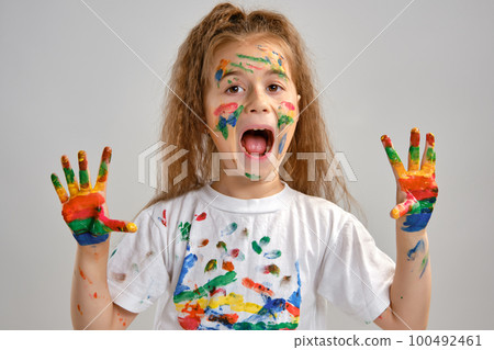 Little girl in white t-shirt is posing standing isolated on white and gesticulating with her painted in different colors palms. Art studio. Close-up. 100492461