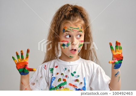 Little girl in white t-shirt is posing standing isolated on white and gesticulating with her painted in different colors palms. Art studio. Close-up. 100492463