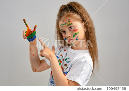 Little girl in white t-shirt is posing standing isolated on white and gesticulating with her painted in different colors palms. Art studio. Close-up. 100492466