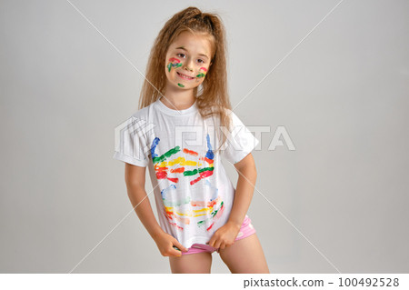 Little girl in white painted t-shirt is posing standing isolated on white and gesticulating with her colored in different paints palms and face. Art studio. Close-up. 100492528