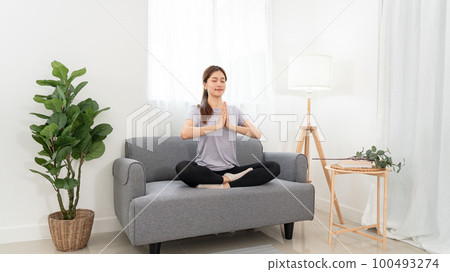 Young woman is relaxing into the lotus position with namaste gesture for meditation on couch Young woman is relaxing into the lotus position with namaste gesture for meditation on couch 100493274