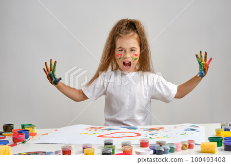Little girl in white t-shirt sitting at table with whatman and paints on it, posing with painted face and hands. Isolated on white. Medium close-up. 100493408