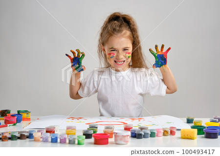 Little girl in white t-shirt sitting at table with whatman and paints on it, posing with painted face and hands. Isolated on white. Medium close-up. 100493417