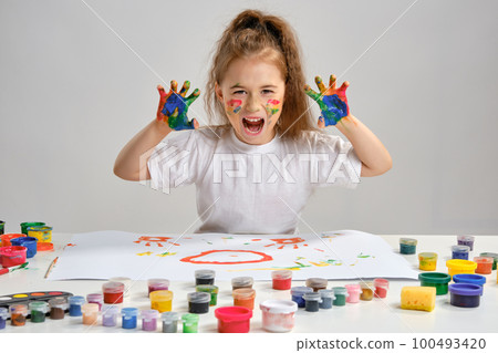Little girl in white t-shirt sitting at table with whatman and paints on it, posing with painted face and hands. Isolated on white. Medium close-up. 100493420