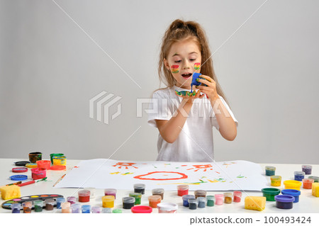 Little girl in white t-shirt sitting at table with whatman and paints on it, posing with painted face and hands. Isolated on white. Medium close-up. 100493424