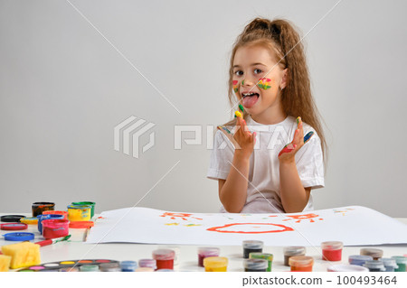Little girl in white t-shirt sitting at table with whatman and paints on it, licking paint from her hands. Isolated on white. Medium close-up. 100493464