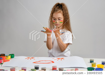 Little girl in white t-shirt sitting at table with whatman and colorful paints, painting on it with her hands. Isolated on white. Medium close-up. 100493505