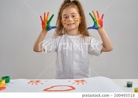 Little girl in white t-shirt sitting at table with whatman and colorful paints, showing her painted hands. Isolated on white. Medium close-up. 100493518
