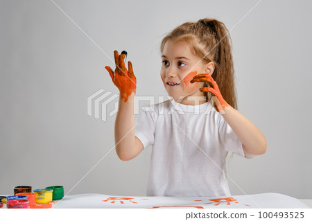 Little girl in white t-shirt sitting at table with whatman and colorful paints on it, painting her hands. Isolated on white. Medium close-up. Little girl in white t-shirt sitting at table with whatman and colorful paints on it, painting her hands. Isolated on white. Medium close-up. 100493525
