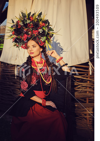 Brunette girl in a black and red embroidered ukrainian authentic national costume and a wreath of flowers is posing against a terrace. Close-up. Brunette girl in a black and red embroidered ukrainian authentic national costume and a wreath of flowers is posing against a terrace. Close-up. 100493589