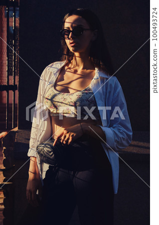 Portrait of a girl in dark sunglasses posing in city near trellised fence. Dressed in top with floral print, white shirt, black trousers, waist bag. Portrait of a girl in dark sunglasses posing in city near trellised fence. Dressed in top with floral print, white shirt, black trousers, waist bag. 100493724