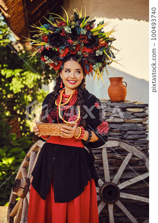 Brunette girl in black and red embroidered ukrainian authentic national costume and a wreath of flowers is posing standing against a white hut. Brunette girl in black and red embroidered ukrainian authentic national costume and a wreath of flowers is posing standing against a white hut. 100493740