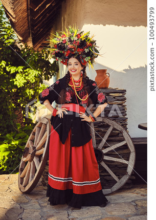 Brunette girl in black and red embroidered ukrainian authentic national costume and a wreath of flowers is posing standing against a white hut. 100493799