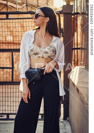 Portrait of a girl in dark sunglasses posing in city against a trellised fence. Dressed in top with floral print, white shirt, black trousers, waist bag. 100493892