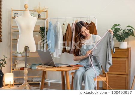 Female online seller is using laptop to broadcast live on Vlog for introduce fashions while measuring 100493937