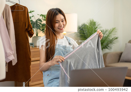 Female online seller is using laptop to broadcast live on Vlog for introduce fashions while measuring 100493942
