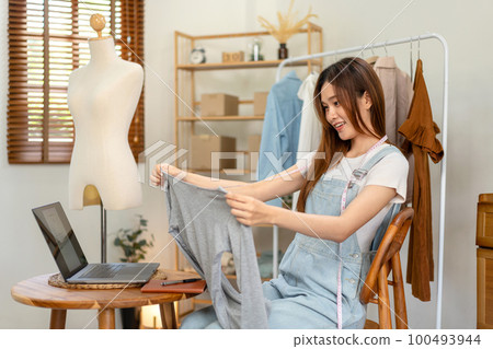Female online seller is using laptop to broadcast live on Vlog for introduce fashions while measuring 100493944