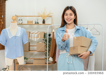 Female online seller is using smartphone to broadcast live on Vlog and holding cardboard boxes while making 100493968