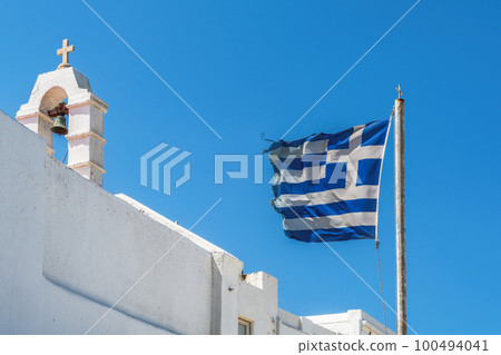 Greek flag waving on a must under strong wind against blue sky background, before a white Orthodox church, with belltower and Christian cross on top at Mykonos Island. Greek flag waving on a must under strong wind against blue sky background, before a white Orthodox church, with belltower and Christian cross on top at Mykonos Island. 100494041