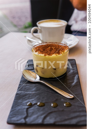 Tiramisu sweet dessert served in Italy with a spoon on a table with a defocused background of a cup of hot cappuccino coffee. Tiramisu sweet dessert served in Italy with a spoon on a table with a defocused background of a cup of hot cappuccino coffee. 100494044