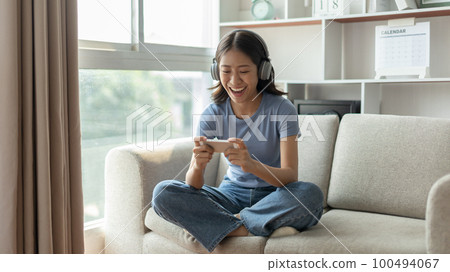 Asian woman playing games on mobile phone and wearing headphones having fun, Play games on the sofa in the living room on weekends, Resting at home, Comfort zone, Touch screen mobile phone. 100494067