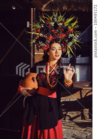 Brunette girl in a black and red ukrainian embroidered authentic national costume and a wreath of flowers is posing standing at the gate. Brunette girl in a black and red ukrainian embroidered authentic national costume and a wreath of flowers is posing standing at the gate. 100495172
