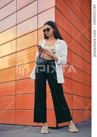 Girl in sunglasses posing in city against an orange building. Dressed in top with floral print, white shirt, black trousers, waist bag and sneakers. Girl in sunglasses posing in city against an orange building. Dressed in top with floral print, white shirt, black trousers, waist bag and sneakers. 100495194