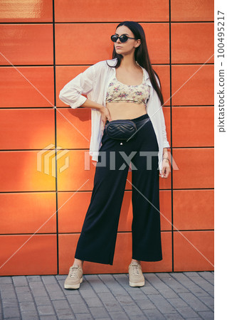 Girl in sunglasses posing in city against an orange building. Dressed in top with floral print, white shirt, black trousers, waist bag and sneakers. 100495217