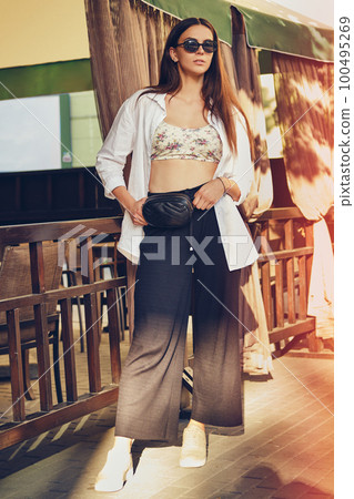 Girl in dark sunglasses posing in city near summer terrace. Dressed in top with floral print, white shirt, black trousers, waist bag and sneakers. 100495269