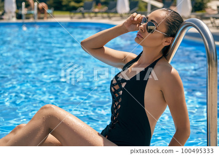 Portrait of a female having rest and posing in a swimming pool. Dressed in a black swimsuit and sunglasses. 100495333