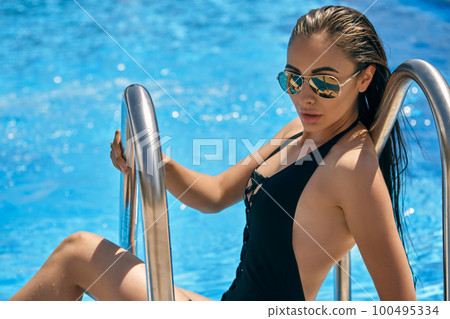 Portrait of a female having rest and posing in a swimming pool. Dressed in a black swimsuit and sunglasses. Portrait of a female having rest and posing in a swimming pool. Dressed in a black swimsuit and sunglasses. 100495334