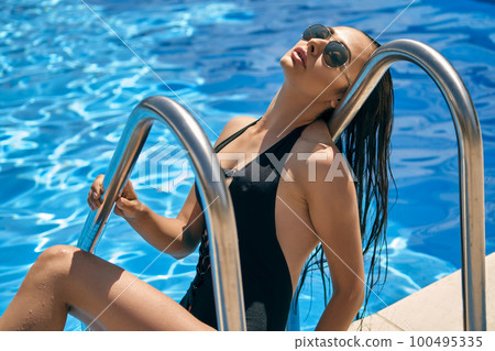 Portrait of a female having rest and posing in a swimming pool. Dressed in a black swimsuit and sunglasses. 100495335