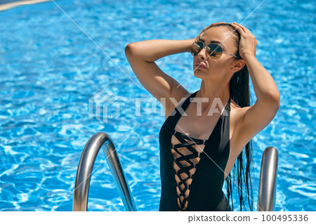 Portrait of a female having rest and posing in a swimming pool. Dressed in a black swimsuit and sunglasses. 100495336