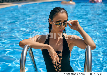Portrait of a female having rest and posing in a swimming pool. Dressed in a black swimsuit and sunglasses. Portrait of a female having rest and posing in a swimming pool. Dressed in a black swimsuit and sunglasses. 100495340