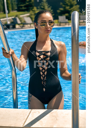 Portrait of a female having rest and posing in a swimming pool. Dressed in a black swimsuit and sunglasses. Portrait of a female having rest and posing in a swimming pool. Dressed in a black swimsuit and sunglasses. 100495368